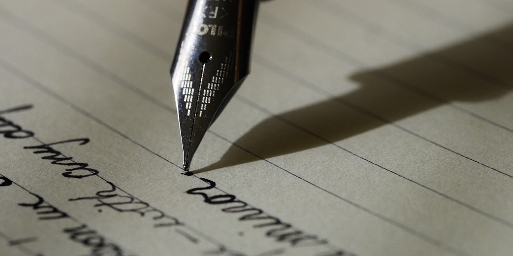 A piece of lined paper with indistinct cursive letters written on it in black ink, two lines have been written and a pen tip is pointed down into the paper, starting a third